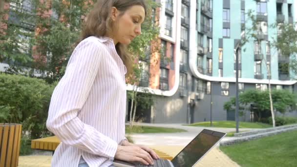 Dizüstü bilgisayarın klavyesinde yazan genç bir kadın, modern binanın arka planında dijital tablet kullanan bir iş kadını, açık havada çalışan bir hesap yöneticisi, uzak bir iş. — Stok video