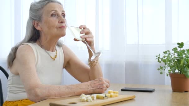 Belle vieille grand-mère aux cheveux gris et visage ridé assis à la table à la maison sur fond de fenêtre avec verre de champagne, fête des mères, retraite heureuse — Video