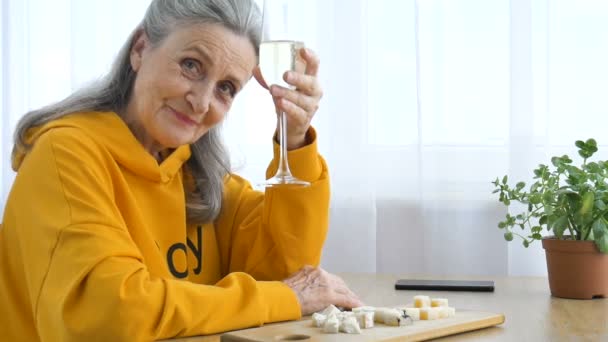 Belle vieille grand-mère aux cheveux gris et visage ridé assis à la table à la maison sur fond de fenêtre avec verre de champagne, fête des mères, retraite heureuse — Video