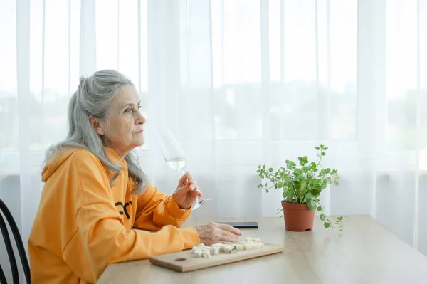 Красивая старая бабушка с седыми волосами и лицом с морщинами, сидя за столом дома на фоне окна с бокалом шампанского, День матери, счастливого выхода на пенсию — стоковое фото