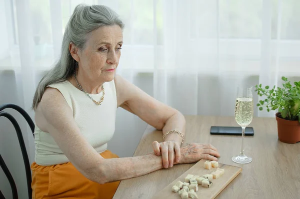 Beautiful old grandmother with grey hair and face with wrinkles sitting at the table at home on window background with glass of champagne, mothers day, happy retirement — Stock Photo, Image
