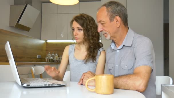魅力的な若い娘と高齢者の父親は、アパートのモダンなキッチンに座ってラップトップを使用しています。女性は家庭で父親のために技術を教えています幸せな退職の概念 — ストック動画