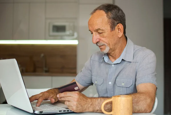 Традиционный зрелый мужчина работает из дома с ноутбуком, сидя за столом на своей кухне, покупая что-то с кредитной картой онлайн, счастливая пенсия, домашние офисные концепции — стоковое фото