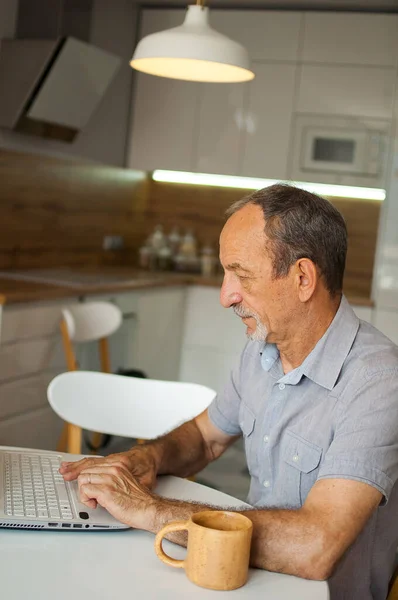 时尚的、成熟的男人在家里工作，他的厨房里坐着笔记本电脑，享受着退休生活，有了家庭办公的理念 — 图库照片