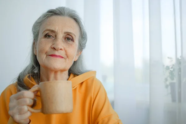 Happy vivacious middle-aged blond woman holding a cup of tea or coffee looking at the camera with a beaming warm smile — Stock Photo, Image