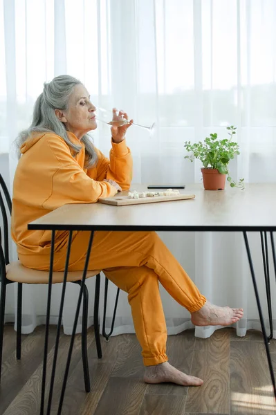 Красивая старая бабушка с седыми волосами и лицом с морщинами, сидя за столом дома на фоне окна с бокалом шампанского, День матери, счастливого выхода на пенсию — стоковое фото