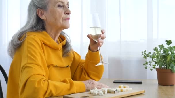 Hermosa abuela de edad con el pelo gris y la cara con arrugas sentado a la mesa en casa en el fondo de la ventana con una copa de champán, día de las madres, feliz jubilación — Vídeos de Stock