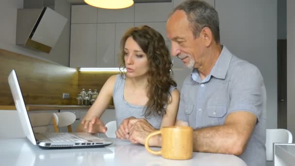 Приваблива молода дочка і старший батько використовують ноутбук, що сидить на сучасній кухні в квартирі. Жінка викладає технології для свого батька вдома, щаслива концепція пенсії — стокове відео