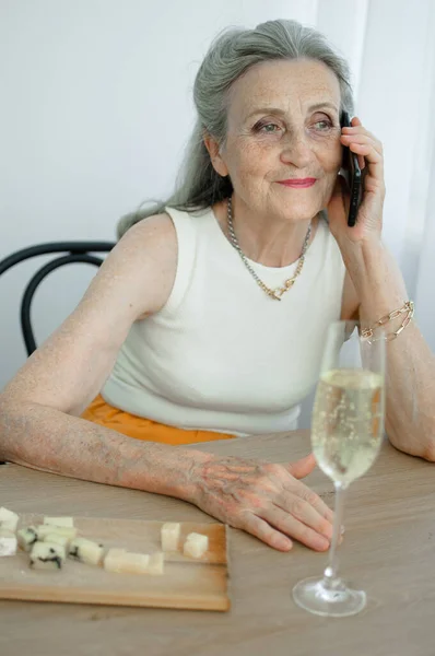 Beautiful old grandmother with grey hair and face with wrinkles is using smartphone, talking with someone and sitting at the table at home on window background, mothers day, happy retirement — Stock Photo, Image