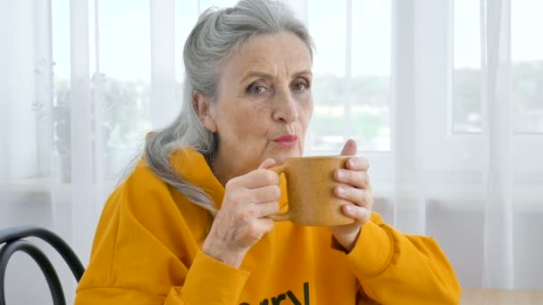 Heureux retraité âgé, excentrique femme aux cheveux gris en jaune boit du thé, regardant pensivement dans une tasse — Video