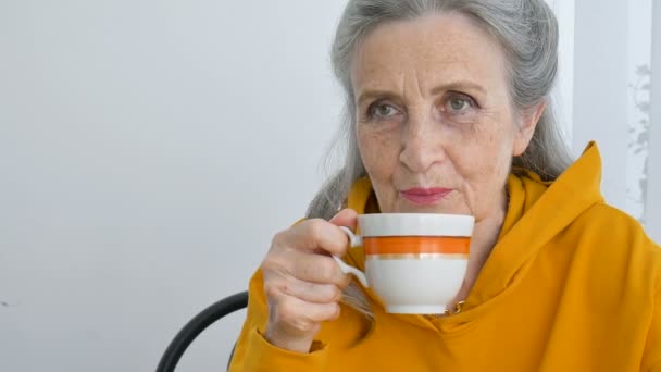 Heureux retraité âgé, excentrique femme aux cheveux gris en jaune boit du thé, regardant pensivement dans une tasse — Video