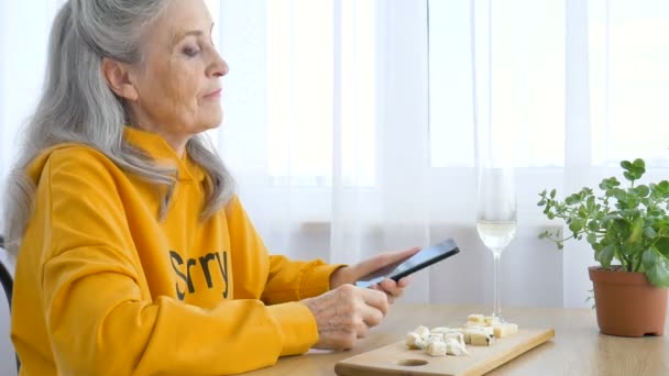 Belle vieille grand-mère avec les cheveux gris et le visage avec des rides utilise smartphone, parler avec quelqu'un et assis à la table à la maison sur fond de fenêtre, fête des mères, retraite heureuse — Video