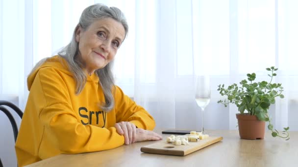 Belle vieille grand-mère aux cheveux gris et visage ridé assis à la table à la maison sur fond de fenêtre avec verre de champagne, fête des mères, retraite heureuse — Video