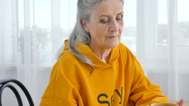 黄色の幸せな高齢者の年金受給者、偏心白髪の女性は、思慮深くマグカップを見てお茶を飲んでいる — ストック動画