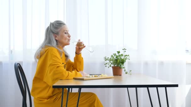 Belle vieille grand-mère aux cheveux gris et visage ridé assis à la table à la maison sur fond de fenêtre avec verre de champagne, fête des mères, retraite heureuse — Video