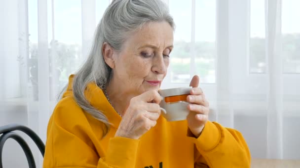 Heureux retraité âgé, excentrique femme aux cheveux gris en jaune boit du thé, regardant pensivement dans une tasse — Video