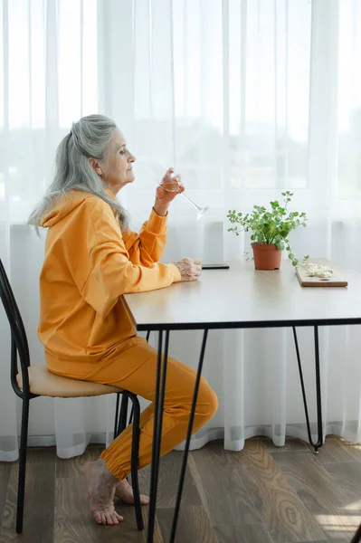 Красивая старая бабушка с седыми волосами и лицо с морщинами с помощью смартфона, разговаривая с кем-то и сидя за столом дома на фоне окна, День матери, счастливый выход на пенсию — стоковое фото