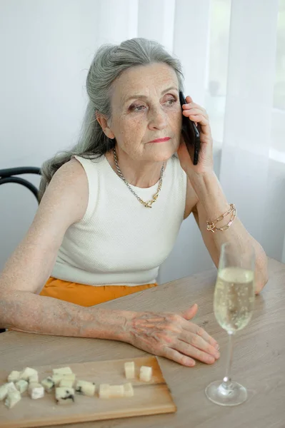 Beautiful old grandmother with grey hair and face with wrinkles is using smartphone, talking with someone and sitting at the table at home on window background, mothers day, happy retirement — Stock Photo, Image