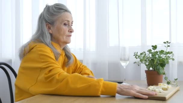 Hermosa abuela de edad con el pelo gris y la cara con arrugas sentado a la mesa en casa en el fondo de la ventana con una copa de champán, día de las madres, feliz jubilación — Vídeo de stock