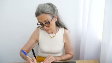 Gözlüklü yaşlı gri saçlı iş kadını masadaki bir kağıda bir şeyler yazıyor. Mutlu emeklilik, istihdam ve iş gücü