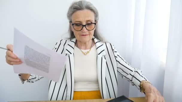 La mujer de negocios de pelo gris mayor con chaqueta a rayas con anteojos está escribiendo algo en un papel aquí, en la oficina sentada en el escritorio. Feliz jubilación, empleo y trabajo — Vídeos de Stock