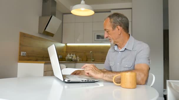 Trendiger, reifer Mann arbeitet von zu Hause aus mit Laptop am Tisch in seiner Küche, kauft online etwas mit Kreditkarte, glücklicher Ruhestand, Home-Office-Konzepte — Stockvideo