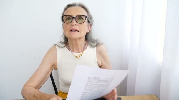 Una mujer de negocios con gafas de pelo gris está escribiendo algo en un papel aquí en la oficina sentada en el escritorio. Feliz jubilación, empleo y trabajo — Vídeo de stock