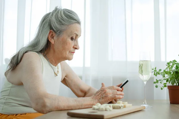 Красивая старая бабушка с седыми волосами и лицо с морщинами с помощью смартфона, разговаривая с кем-то и сидя за столом дома на фоне окна, День матери, счастливый выход на пенсию — стоковое фото