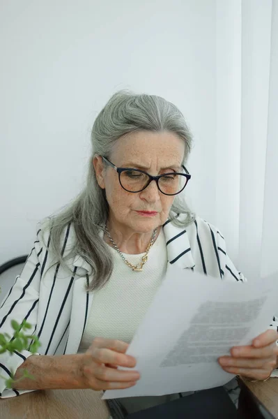 Старшая седовласая деловая женщина в полосатой куртке в очках работает с документами здесь, в офисе, сидя за столом. Счастливого выхода на пенсию, работы и труда — стоковое фото
