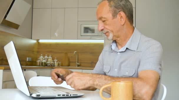 Trendiger, reifer Mann arbeitet von zu Hause aus mit Laptop am Tisch in seiner Küche, kauft online etwas mit Kreditkarte, glücklicher Ruhestand, Home-Office-Konzepte — Stockvideo