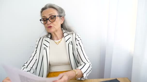 L'anziana donna d'affari dai capelli grigi con giacca a righe e occhiali sta scrivendo qualcosa su un foglio qui dentro, in ufficio, seduta alla scrivania. Buon pensionamento, occupazione e lavoro — Video Stock