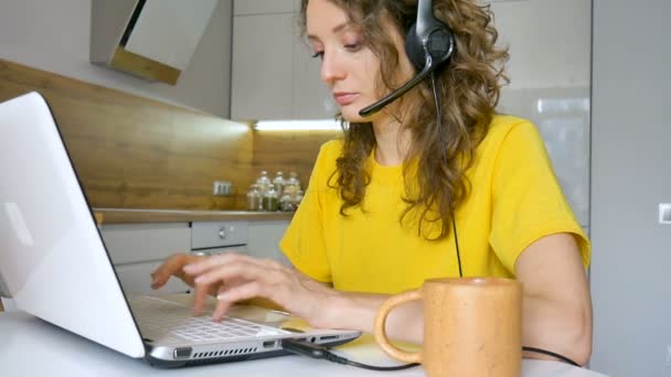 Молодая женщина в ярко-желтой футболке печатает на клавиатуре ноутбука, деловая женщина, использующая цифровой планшет дома на своей кухне, менеджер по работе с клиентами, удаленная работа — стоковое видео