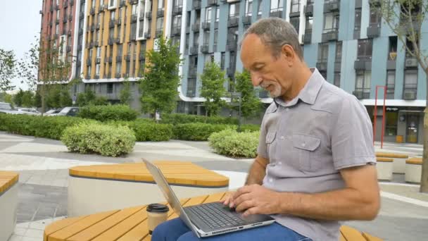 Trendy dojrzały mężczyzna pracuje na zewnątrz z laptopem siedzi na ławce w pobliżu nowego nowoczesnego kompleksu mieszkalnego, szczęśliwych koncepcji emerytalnych — Wideo stockowe