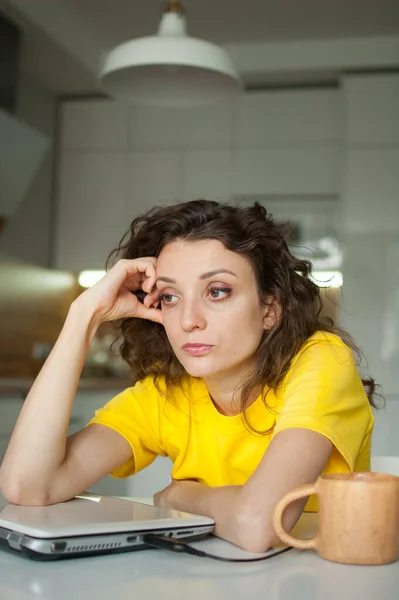 stock image Tired young woman with curly hair and yellow shirt is working from home using her laptop at the kitchen table in her apartment, remote work, freelance, burnout syndrome