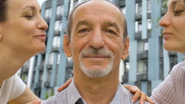 Family portrait of senior father and two adult daughters kissing him outdoors on modern building of new residential complex, fathers day celebration concept — Stock Video