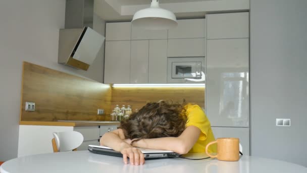 Giovane donna stanca con i capelli ricci e camicia gialla sta lavorando da casa utilizzando il suo computer portatile al tavolo della cucina nel suo appartamento, lavoro a distanza, freelance, sindrome di burnout — Video Stock