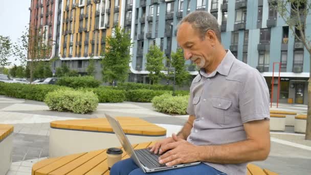Trendiger, reifer Mann arbeitet draußen mit Laptop auf der Bank in der Nähe des neuen modernen Wohnkomplexes, glückliche Rentenkonzepte — Stockvideo