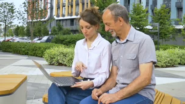 Привлекательная молодая женщина и старший мужчина пользуются ноутбуком, сидя на скамейке в современном жилом комплексе. Случайная встреча вне офиса. Дочь преподает технологию своему отцу — стоковое видео