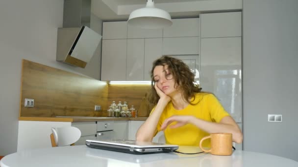 Jeune femme fatiguée avec les cheveux bouclés et chemise jaune travaille de la maison en utilisant son ordinateur portable à la table de la cuisine dans son appartement, travail à distance, freelance, syndrome d'épuisement professionnel — Video