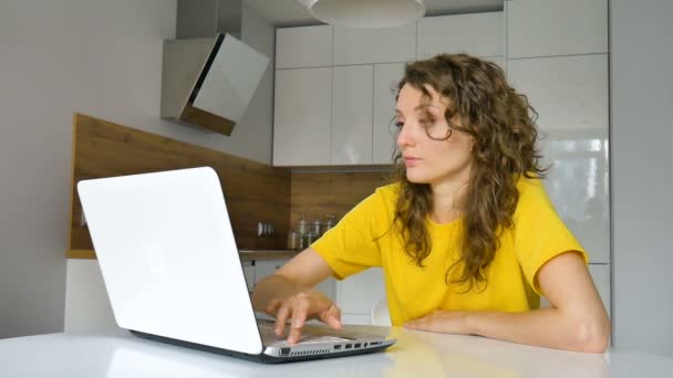 Młoda kobieta w kręconych włosach i żółtej koszuli pracuje z domu za pomocą laptopa przy kuchennym stole w mieszkaniu, zdalna praca, freelance — Wideo stockowe