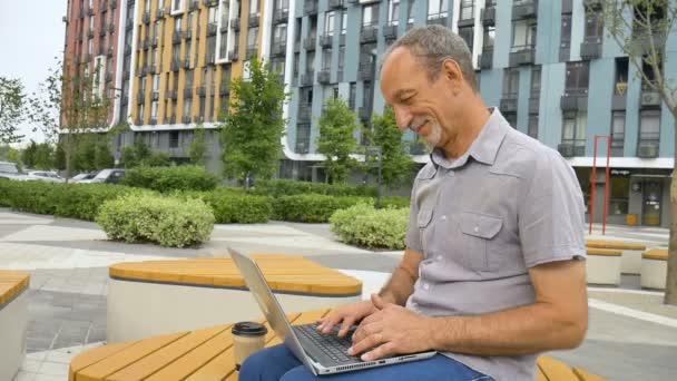 Trendy érett férfi dolgozik kívül laptop ül a padon közelében új modern lakópark, boldog nyugdíjazás fogalmak — Stock videók