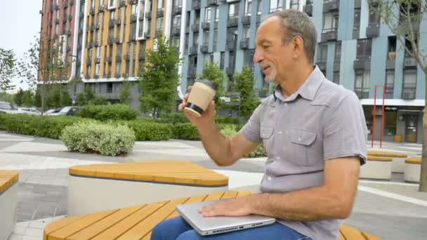Модный зрелый мужчина работает на улице с ноутбуком сидя на скамейке рядом с новым современным жилым комплексом, счастливый пенсионных концепций — стоковое видео