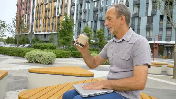 Trendiger, reifer Mann arbeitet draußen mit Laptop auf der Bank in der Nähe des neuen modernen Wohnkomplexes, glückliche Rentenkonzepte — Stockvideo