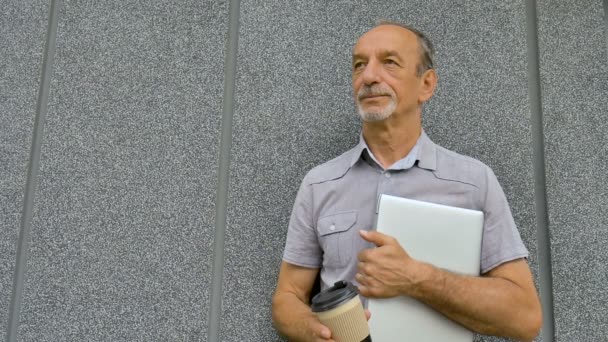 Manligt porträtt av äldre medarbetare med silver laptop och kopp kaffe väntar på ett möte nära mörkgrå vägg på morgonen, distansarbete, kaffepaus — Stockvideo