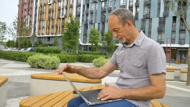 Trendy érett férfi dolgozik kívül laptop ül a padon közelében új modern lakópark, boldog nyugdíjazás fogalmak — Stock videók