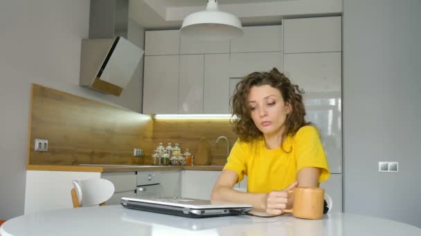 Jovem cansada com cabelo encaracolado e camisa amarela está trabalhando em casa usando seu laptop na mesa da cozinha em seu apartamento, trabalho remoto, freelance, síndrome de burnout — Vídeo de Stock