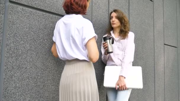 Deux femmes d'affaires prospères parlent d'un projet tout en se tenant près du mur à l'extérieur avec un ordinateur portable pendant une pause de travail — Video