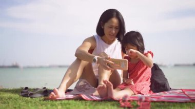 Ağır çekimde Asya kökenli Çinli anne ve kız çocuğu açık deniz pikniğinde akıllı telefona yakından bakıyor.