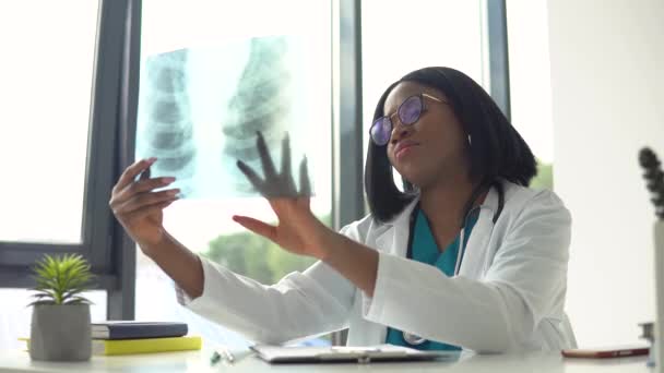 Afrikanische Ärztin mit Brille betrachtet Röntgenbild der Lungen, während sie am Tisch in einer modernen Klinik sitzt — Stockvideo