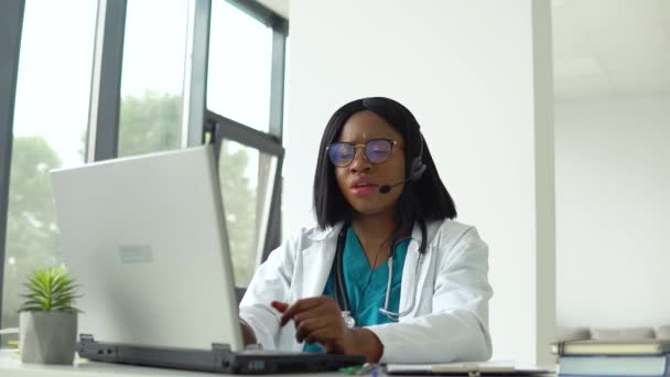 Professional african american female doctor in white medical coat and headset making conference call on laptop computer, consulting distance patient online in video chat — Stock Video